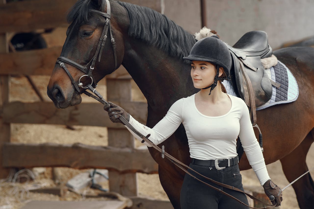 A young woman equestrian in a white blouse stands beside a brown horse, outdoors.